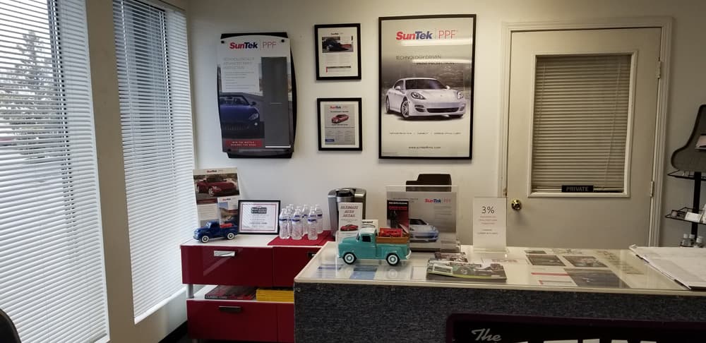A reception area features framed SunTek PPF posters and brochures, a red cabinet with water bottles and car models, and a countertop displaying car-related literature and small Toyotas, indicating a professional auto detailing business specializing in car protection films.